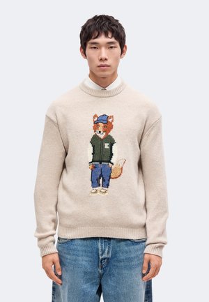 Un homme porte un pull crème orné d’un renard brodé portant une casquette et une veste, avec un jean bleu, sur un fond uni.