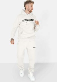 Lichtbeige hoodie en sweatpants set met "SIXTHJUNE" branding. Het materiaal is zacht, met een voorzak en geribbelde manchetten. Witte sneakers.