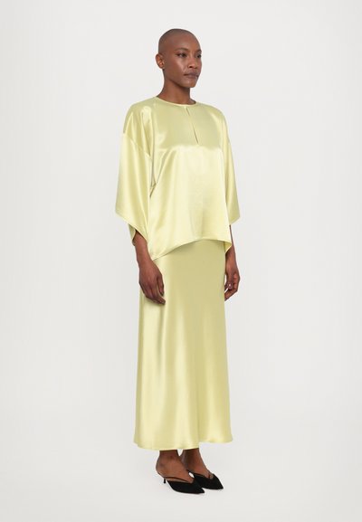 Top de satén amarillo claro con mangas anchas y escote redondo, combinado con una falda larga a juego. Tacones negros de punta afilada completan el atuendo.