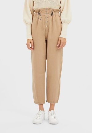 Mujer vistiendo pantalones paperbag de talle alto color beige con botones, combinados con un suéter crema y zapatillas blancas sobre un fondo liso.