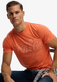 Camiseta de algodón naranja con cuello redondo, mangas cortas y texto impreso en blanco. Se lleva con vaqueros azules y una camisa de cuadros anudada a la cintura.
