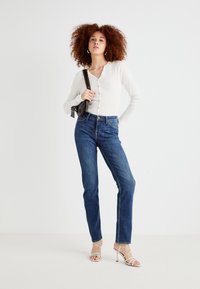 Maglietta bianca a maniche lunghe a coste con bottoni, jeans blu a vita alta e tacchi beige con cinturini, caratterizzati da una vestibilità slim e una texture morbida.