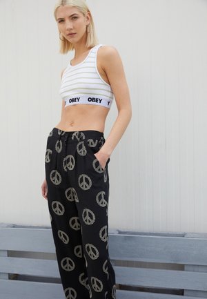 Pantalones negros con un patrón de símbolos de paz, combinados con una camiseta sin mangas blanca y recortada con rayas horizontales y el logo "OBEY" en la banda.