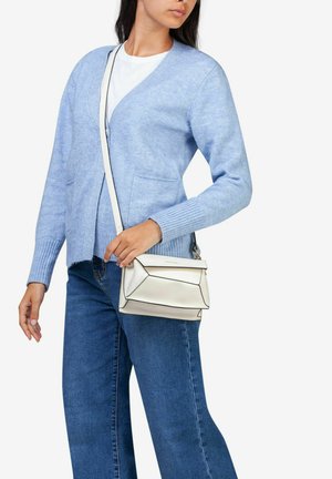 Jasnoniebieski kardigan, biały T-shirt, dżinsy z szerokimi nogawkami. Trzyma małą białą skórzaną torebkę z czarnymi akcentami i paskiem.