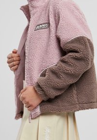 Chaqueta de forro polar en rosa y marrón, con cremallera frontal, cuello alto y detalle de ribete blanco. Parche de logo en el pecho. Textura suave.