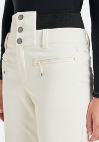 Crème-kleurige broek met hoge taille, drie drukknoopjes en een zwarte elastische tailleband. Bevat twee met een rits afsluitbare zakken met lipjes.