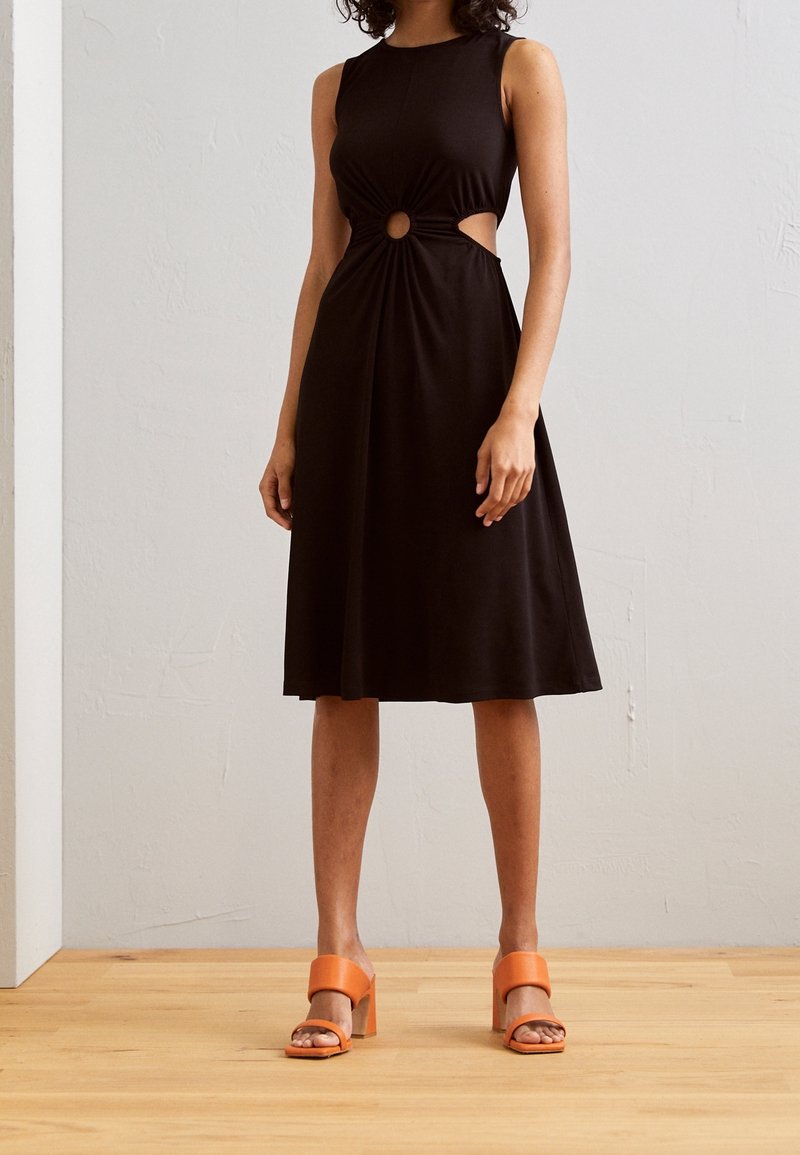 Robe noire sans manches avec détail noué et découpe circulaire à la taille, associée à des sandales à talons blocs orange. Tissu lisse.