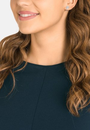 Silver stud earrings with a geometric design, worn with a dark teal top. Hair is wavy and shoulder-length, framing the face neatly.