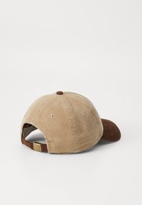 Corduroy baseball cap in beige and brown, with a curved visor, textured fabric, adjustable strap, and a small logo detail on the back.
