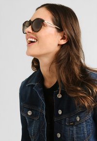 Óculos de sol com lentes escuras e armações redondas, com um realce dourado nas hastes. Usados com uma jaqueta de ganga azul com botões prateados.
