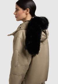 Šviesiai rudos spalvos pūkinė striukė su nuimamu juodu dirbtinio kailio apykakle. Turi užtrauktuką, gobtuvą ir kišenę ant rankovės. Lygi tekstūra.