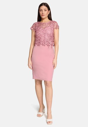 Vrouw staand in een knielange roze jurk met een bovenkant van bloemige kant en kapmouwtjes, gecombineerd met witte open-toe sandalen met blokhak.