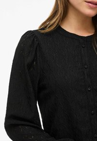 Czarna bluzka wykonana z fakturowanej tkaniny z wzorem diamentowym, z okrągłym dekoltem, zapinana na guziki, z długimi bufiastymi rękawami.
