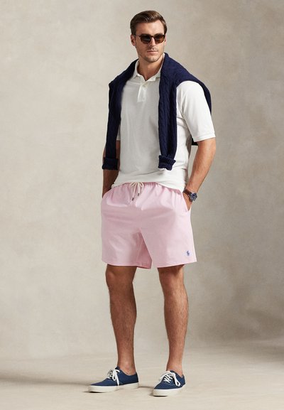 Bărbat îmbrăcat cu tricou polo alb, pantaloni scurți roz, adidași bleumarin, ochelari de soare și pulover bleumarin așezat pe umeri, stând cu mâinile în buzunare.