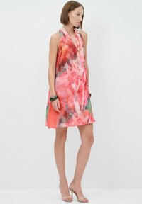 Robe sans manches dans un motif floral aquarelle vibrant rose, rouge et vert ; tissu léger ; tombe au-dessus du genou ; associée à des talons à lanières.