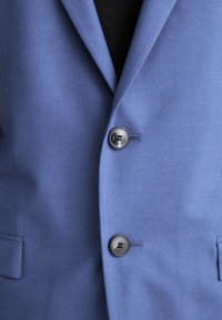 Sacou bleu deschis cu o textură fină, dotat cu două butoane negre, guler cu tăietură și buzunare laterale. Croială ajustată.