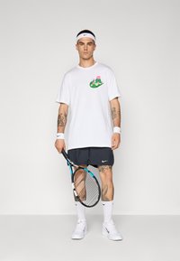 Camiseta blanca de manga corta con un gráfico floral, combinada con pantalones cortos oscuros. Los accesorios incluyen una cinta para la cabeza blanca y muñequeras, sosteniendo una raqueta de tenis.