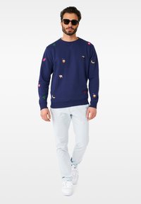 Sudadera azul marino con diversos motivos bordados, incluyendo frutas y animales. Combinada con pantalones azul claro y zapatillas blancas.
