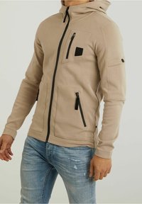 Felpa beige con zip frontale, zip nere a contrasto e due tasche anteriori. Presenta un cappuccio e una toppa sul petto. Tessuto liscio.