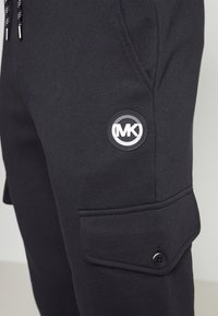 Zbliżenie czarnej nogawki spodni cargos z przednią kieszenią, klapką na guzik, sznurkiem i okrągłą łatą z logo marki MK.