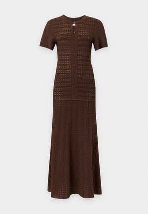 Femme aux cheveux bouclés portant une robe maxi marron foncé à manches courtes boutonnée, avec des perforations décoratives et des talons ouverts noirs.