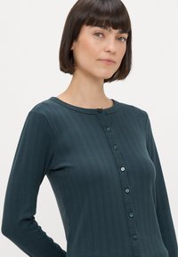Ciemnozielona bluzka z długim rękawem wykonana z prążkowanego materiału, z okrągłym dekoltem, zapięciem na guziki z przodu oraz subtelnym wzorem teksturowym.