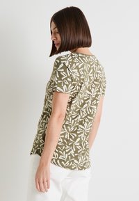 Top de manga corta verde oliva con un patrón de hojas blancas, textura suave, cuello redondo y un dobladillo ligeramente curvado.