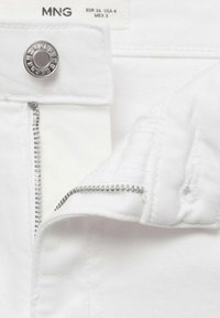 Gros plan sur un pantalon blanc dézippé avec un bouton et une fermeture éclair argentés, montrant l'étiquette de la marque et les détails de la taille à l'intérieur de la ceinture.