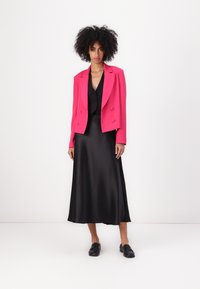 Fuchsia dubbelgebreide blazer over een zwarte satijnen midi-jurk, aangevuld met zwarte loafers. Strakke texturen en gedurfde kleurcontrasten.