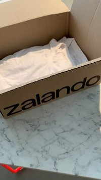 Brązowy kartonowy pudełko z logo Zalando, zawierające złożoną białą koszulkę, leży na marmurowej powierzchni z widocznymi wzorami.