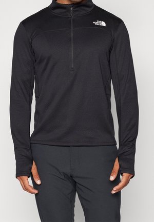 Homme portant un pull-over North Face noir à manches longues avec une fermeture éclair à moitié et des trous pour les pouces dans les manches.