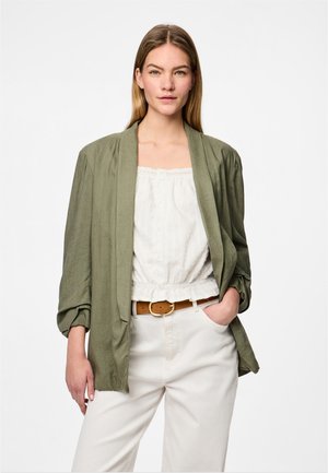 Vrouw met stijl blond haar draagt een olijfgroene blazer over een witte blouse en witte broek met een bruine riem, staand met één hand in de zak.