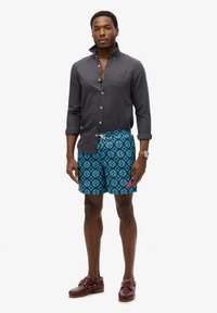 Kortearm overhemd in donkergrijs, gecombineerd met blauwe zwembroek met een geometrisch patroon en rode accenten. Bruine bootschoenen.