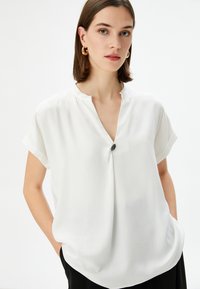 Blusa bianca a maniche corte con colletto a revers, caratterizzata da un dettaglio con bottone centrale. Realizzata in tessuto morbido con una vestibilità ampia.