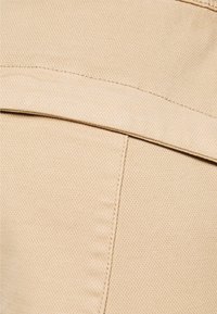 Beige stof met een geweven structuur, met zichtbare stiksels en een zaknaad. Glad oppervlak met een subtiele glans.
