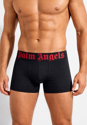 Torso maschile che indossa boxer neri con logo rosso "Palm Angels" sulla cintura, mostrando muscoli addominali definiti e braccia ai lati.