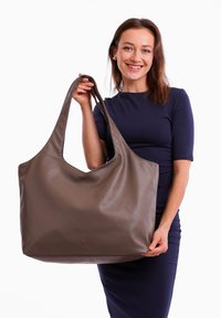 Brown leather tote bag with a smooth texture, large slouchy design, and wide shoulder straps, held by a person wearing a navy dress.