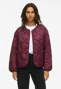 Tânără cu păr creț, purtând o jachetă bordeaux matlasată peste o cămașă albă și pantaloni negri, stând pe un fundal alb.