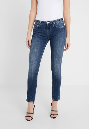 Vrouw draagt donkerblauwe skinny jeans met vervaagde textuur, gecombineerd met zwarte sandaaltjes met hoge hakken en bandjes en een witte mouwloze top.