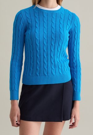 Nainen, joka pukeutunut kirkkaan siniseen palmikkoneulepuseroon valkoisen paidan päällä ja mustaan hameeseen, seisoo yksinkertaisen taustan edessä.