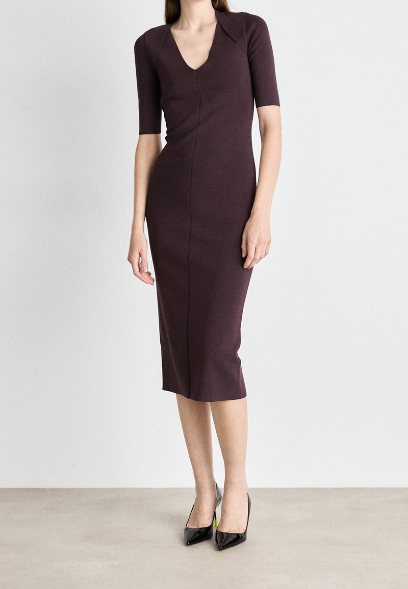 Femme portant une robe ajustée, longueur genou, de couleur violet foncé avec des manches courtes et des talons hauts noirs à bout pointu, debout contre un mur uni.