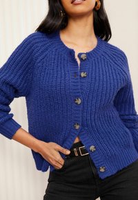 Mėlynas mezginys kardiganas su raukšlėtu tekstūru, apvaliu iškirpimu ir šešiais dideliais mygtukais priekyje. Trumpas ilgis, ilgos rankovės, laisvas siluetas.