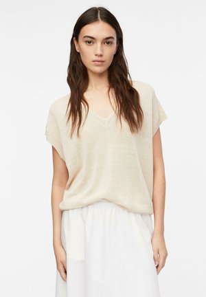 Giovane donna con lunghi capelli castani, indossa una maglietta beige a maniche corte e una gonna bianca, in piedi davanti a uno sfondo semplice.