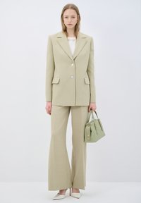 Lichtgroen pak met een getailleerde, dubbelgebreide blazer en wijde broekspijpen. Vergezeld van een kleine groene handtas en witte hakken.