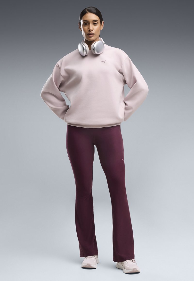 Femme debout les mains sur les hanches, portant un sweat-shirt rose pâle, un legging bordeaux, des baskets blanches et un casque argenté autour du cou.