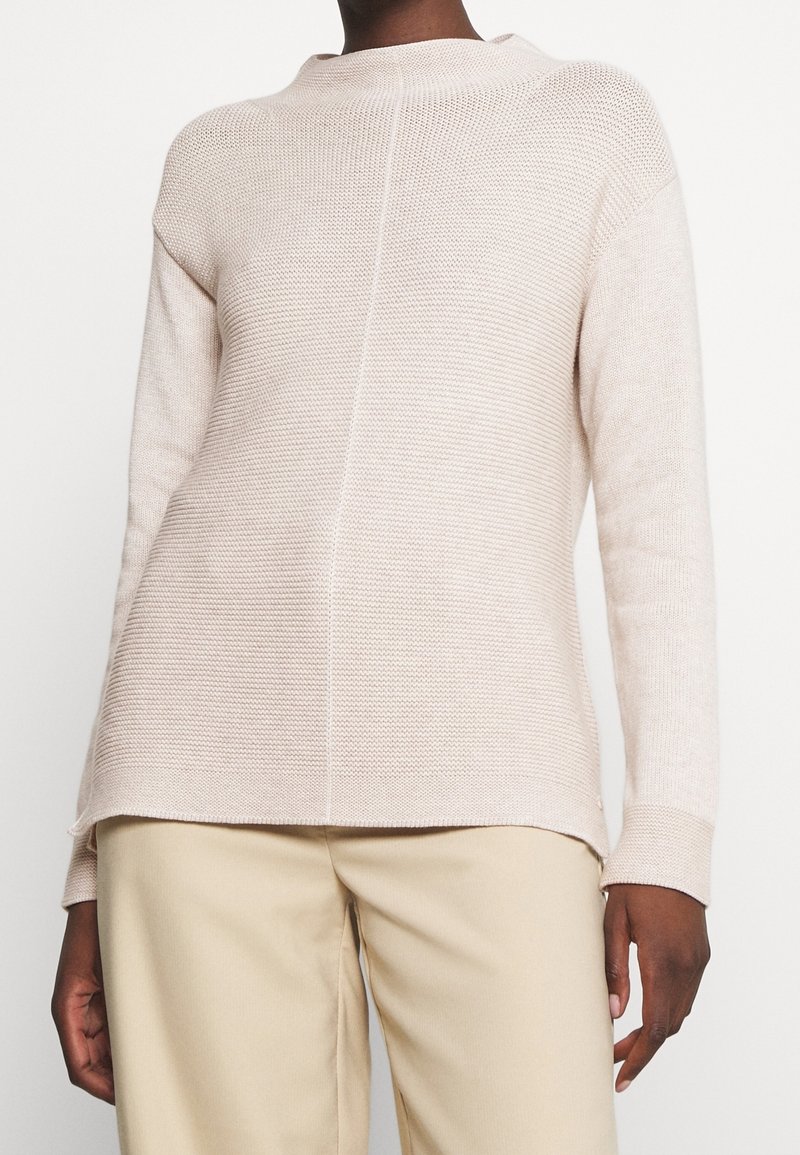 Personne portant un pull en maille beige clair texturé à manches longues et un pantalon beige, montré du cou aux hanches.