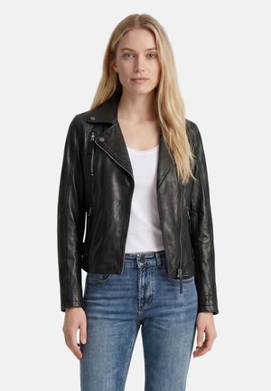 Jeune femme aux longs cheveux blonds portant une veste en cuir noire, une chemise blanche et un jean bleu, debout devant un fond uni.