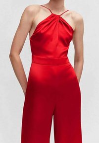 Combinaison salopette en satin rouge à encolure halter avec un détail noué à l'avant, jambes larges et ceinture élastique. Texture lisse et couleur vibrante.