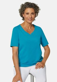 Türkises T-Shirt mit kurzen Ärmeln und V-Ausschnitt, verziert mit dekorativen Strasssteinen am Kragen; getragen zu weißen Hosen. Glatter, dehnbarer Stoff.