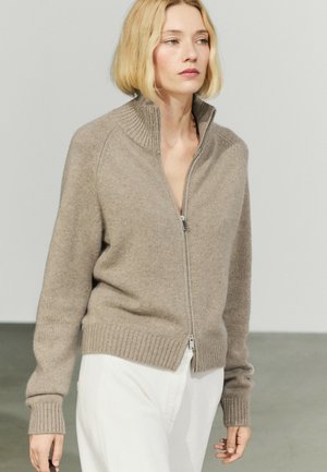Femme aux cheveux blonds courts portant un pull beige zippé et un pantalon blanc, debout devant un fond clair uni.